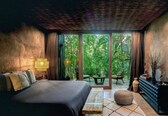 A bedroom with a bed, a nightstand, a lamp, a window, and a view of a garden.