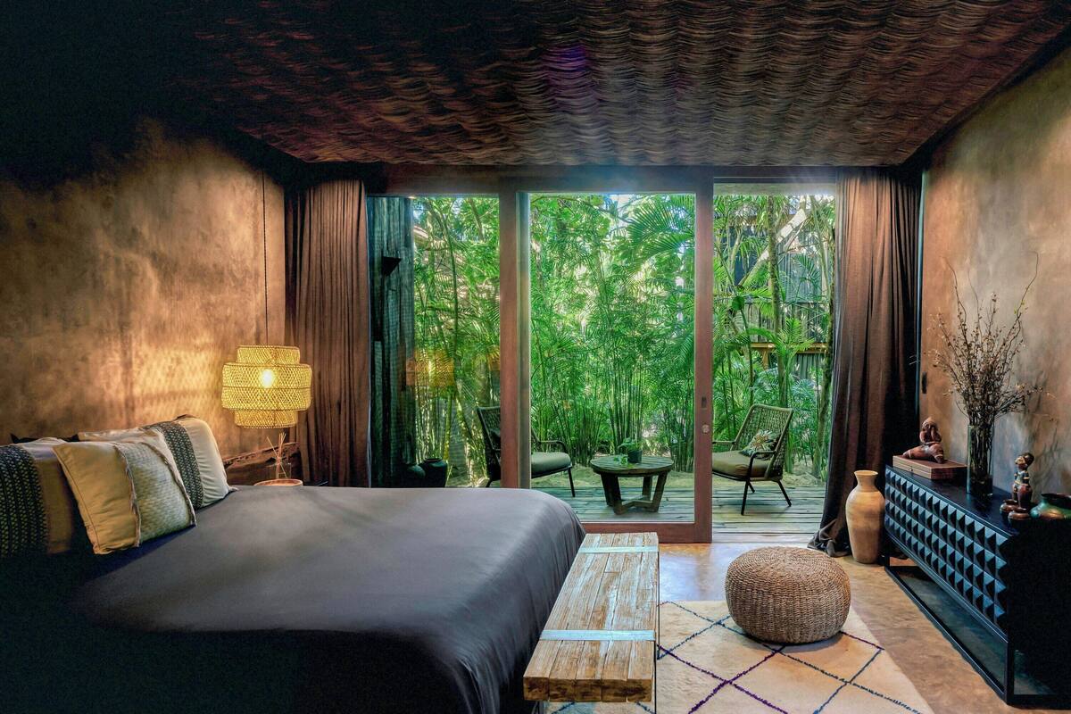 A bedroom with a bed, a nightstand, a lamp, a window, and a view of a garden.