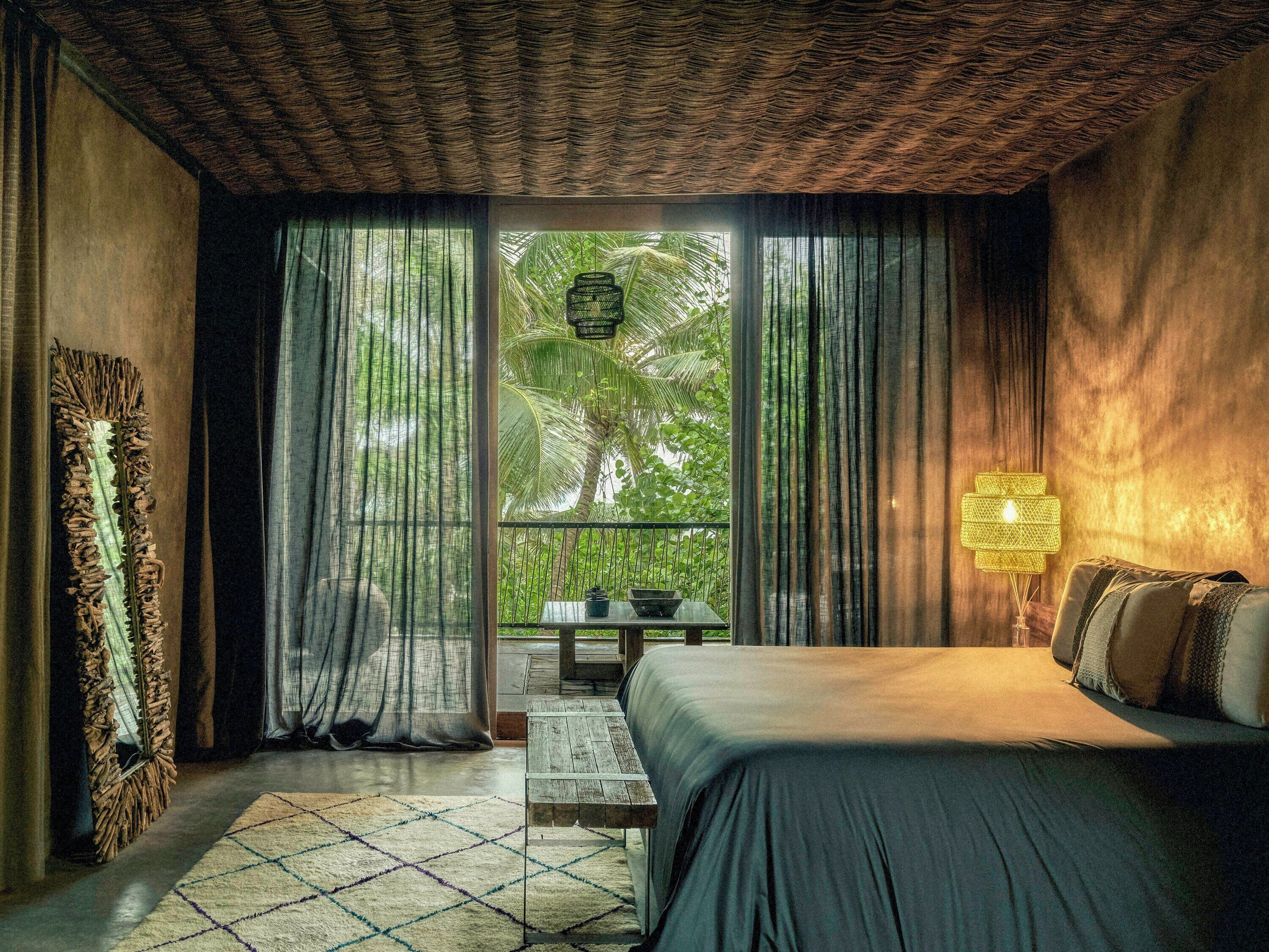 A bedroom with a bed, a nightstand, a lamp, a window with curtains, and a view of greenery outside.