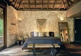 A bedroom with a stone wall, a wooden bed, a thatched roof, and a large mirror.