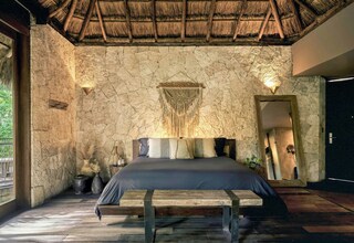 A bedroom with a stone wall, a wooden bed, a thatched roof, and a large mirror.