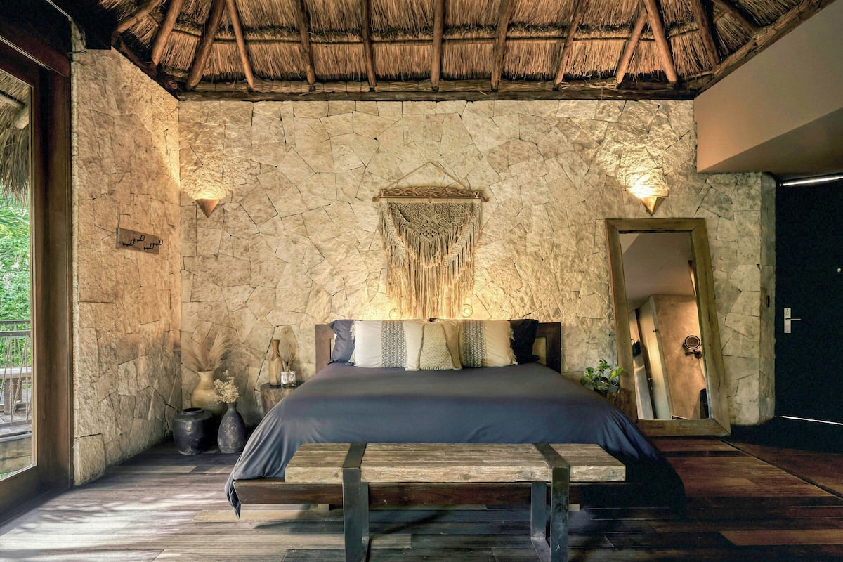 A bedroom with a stone wall, a wooden bed, a thatched roof, and a large mirror.