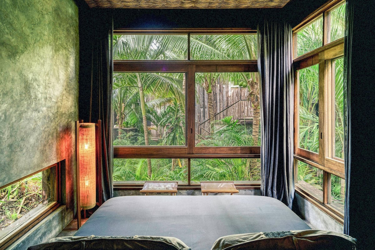 A room with a large window, a bed, a small table, and a lamp.