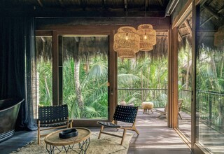 A room with a large glass door leading to a tropical garden, two wicker chairs, a round wooden table, and a hanging light fixture.