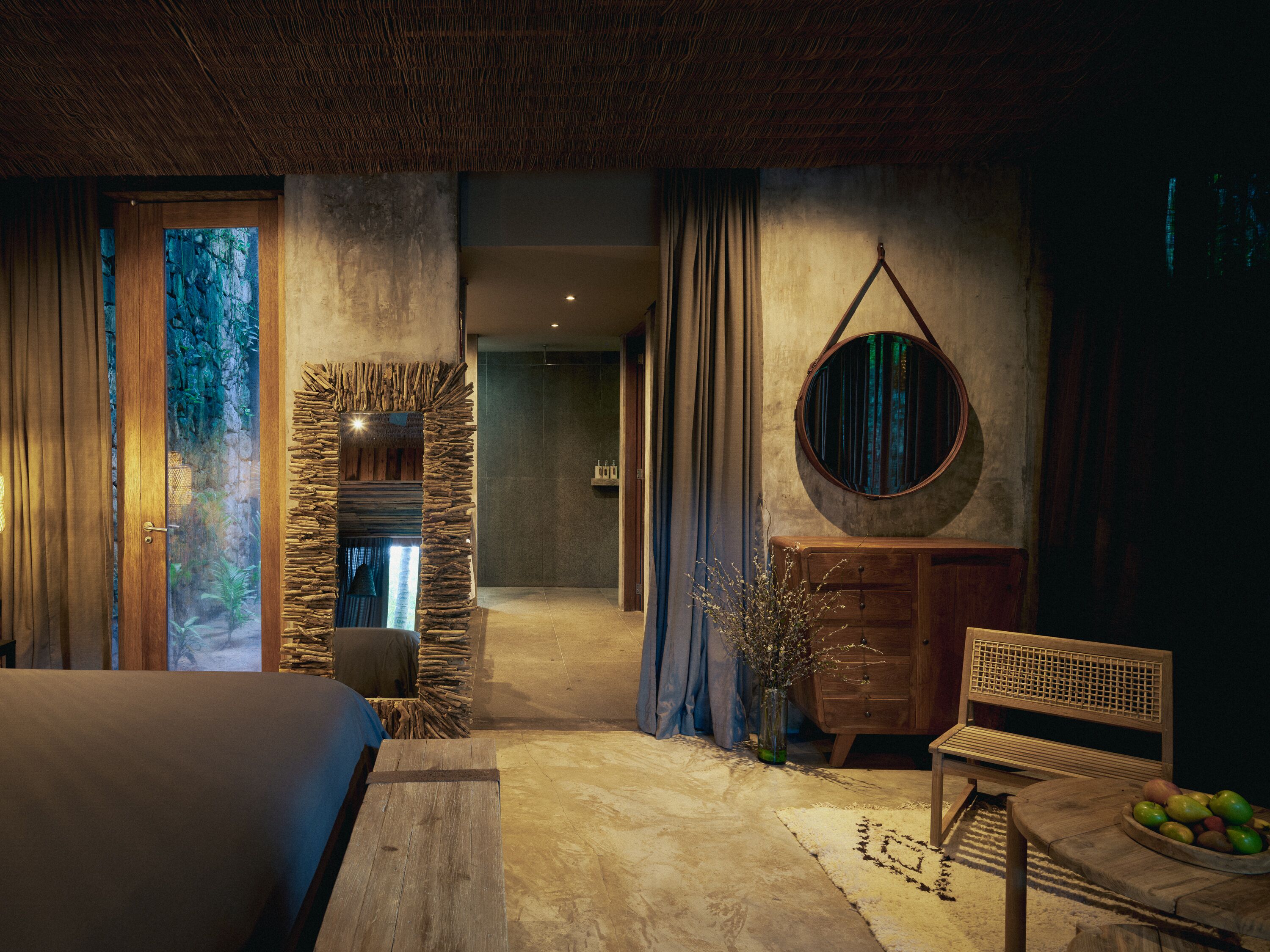 A room with a bed, a wooden dresser, a round mirror, and a stone wall.
