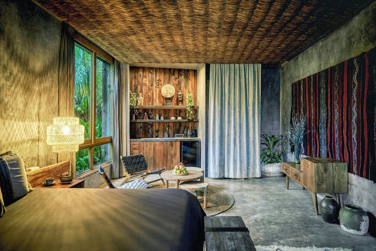 A room with a bed, a desk, a chair, a bookshelf, and a large window with greenery outside.