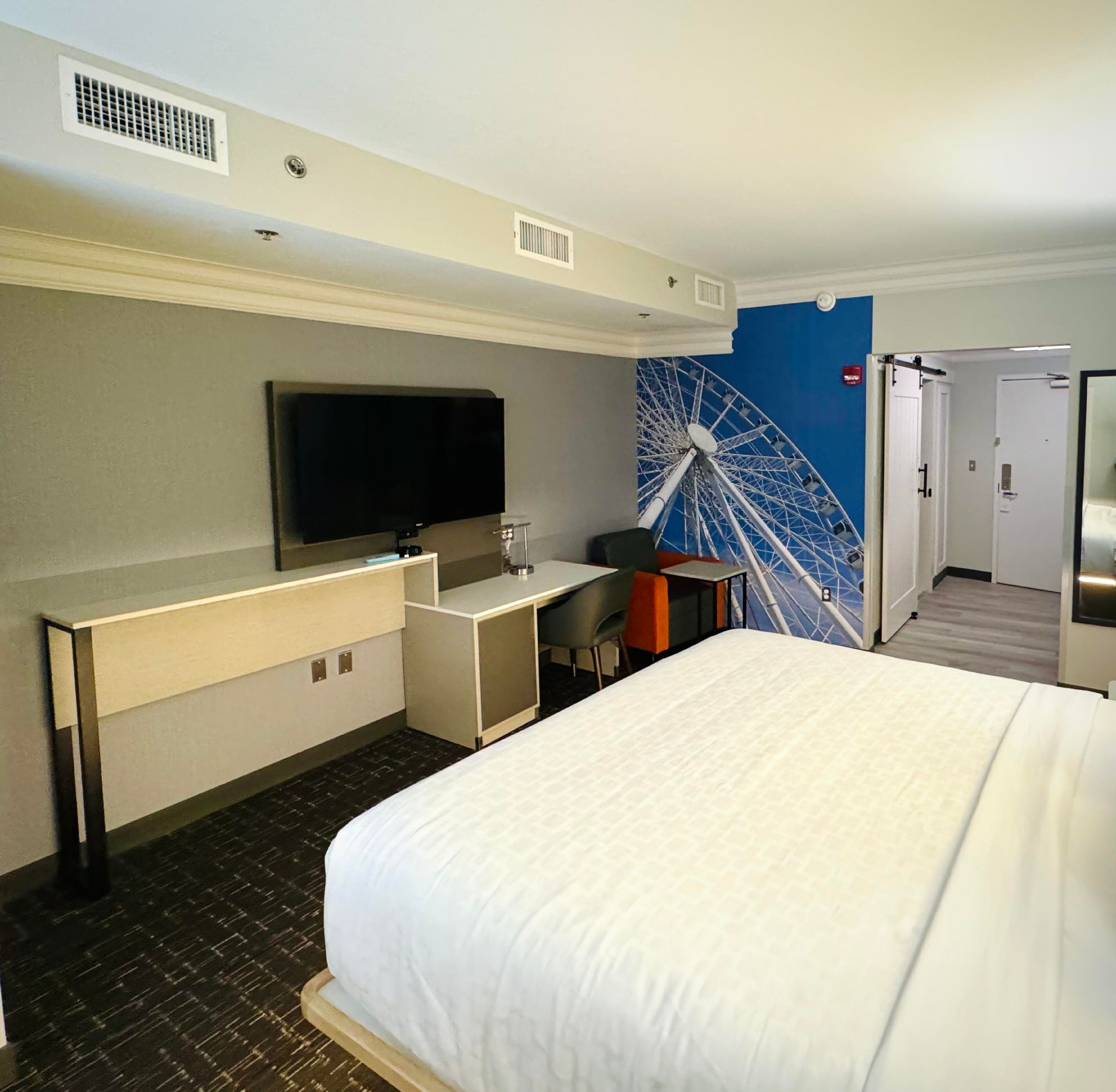 A hotel room with a bed, a TV, a desk, and a wall mural.