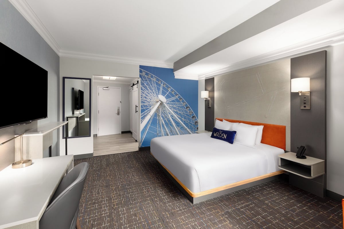 A hotel room with a bed, a TV, a desk, a wall-mounted lamp, and a large wall decoration.