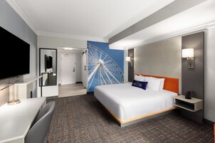 A hotel room with a bed, a TV, a desk, a wall-mounted lamp, and a large wall decoration.