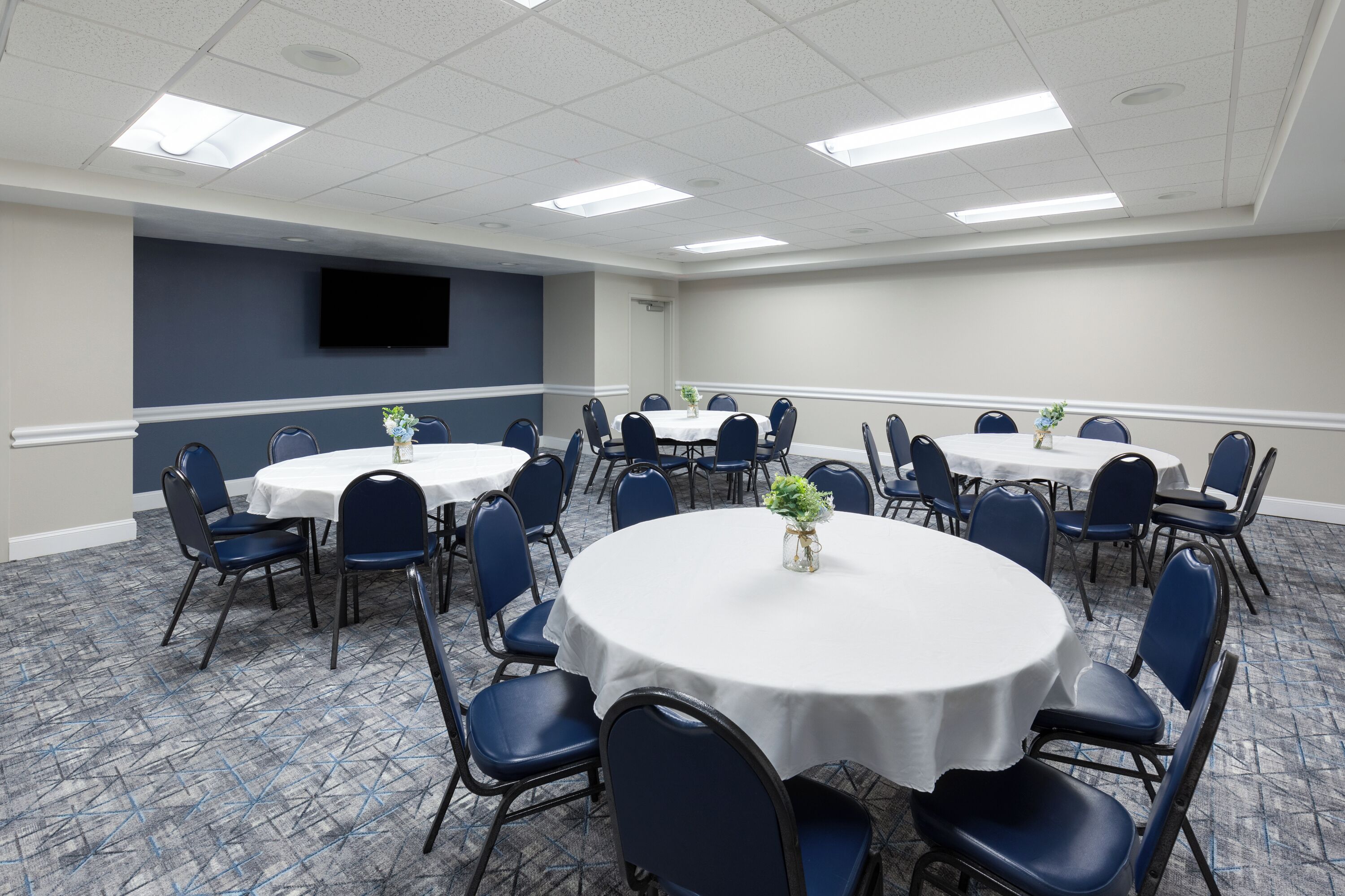Une salle de conférence avec des tables rondes, des chaises bleues et un téléviseur à écran plat fixé au mur.