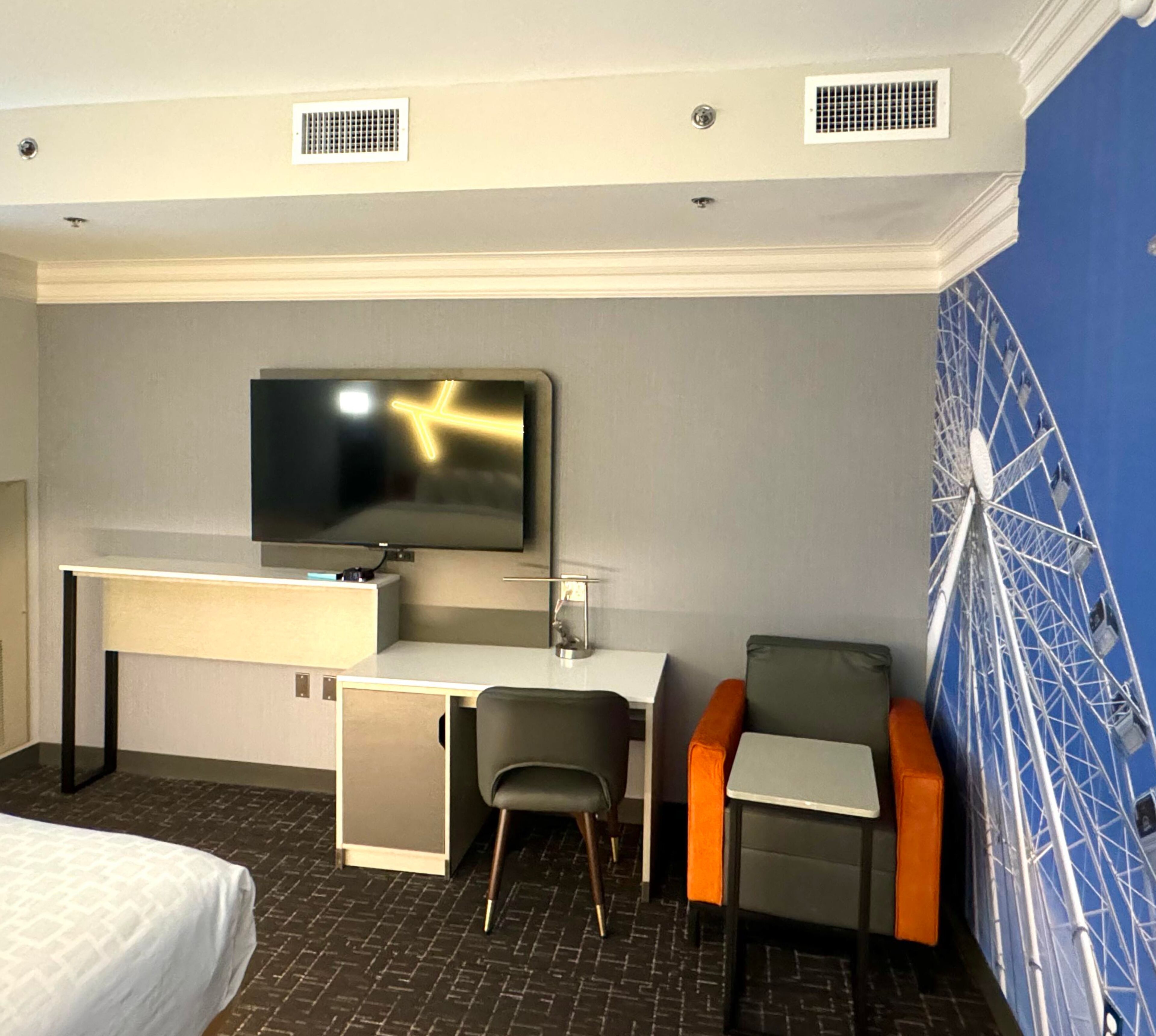 Quarto de hotel com cama, secretária com televisão, duas cadeiras e decoração na parede.