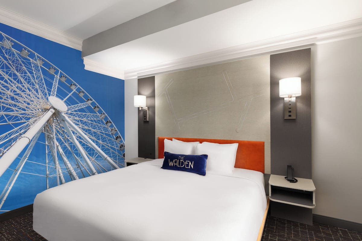 A hotel room with a large bed, a headboard with a name pillow, two bedside lamps, and a wall-mounted shelf.