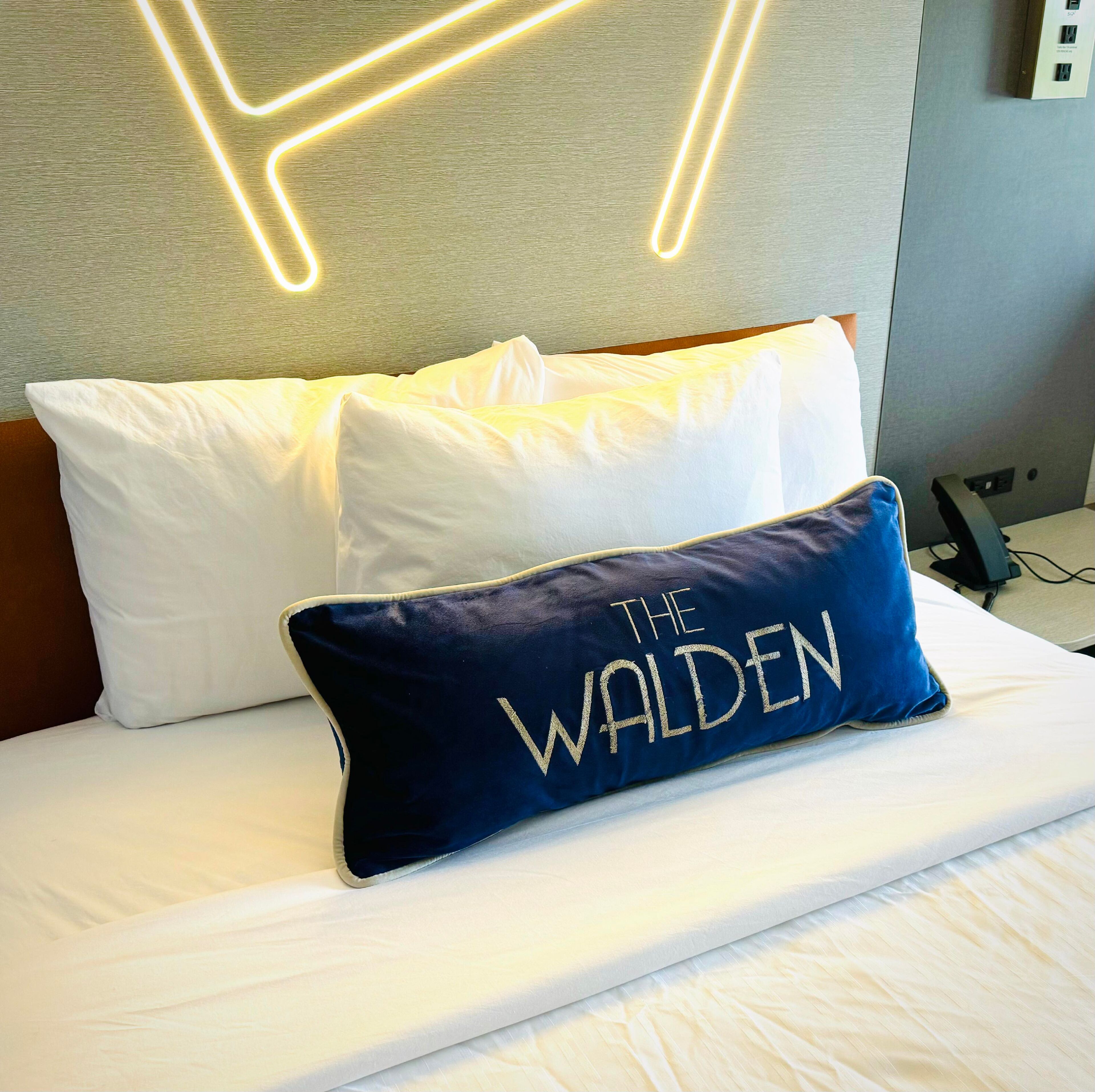 A neatly made bed with white bedding and a dark blue pillow with "THE WALDEN" embroidered on it.