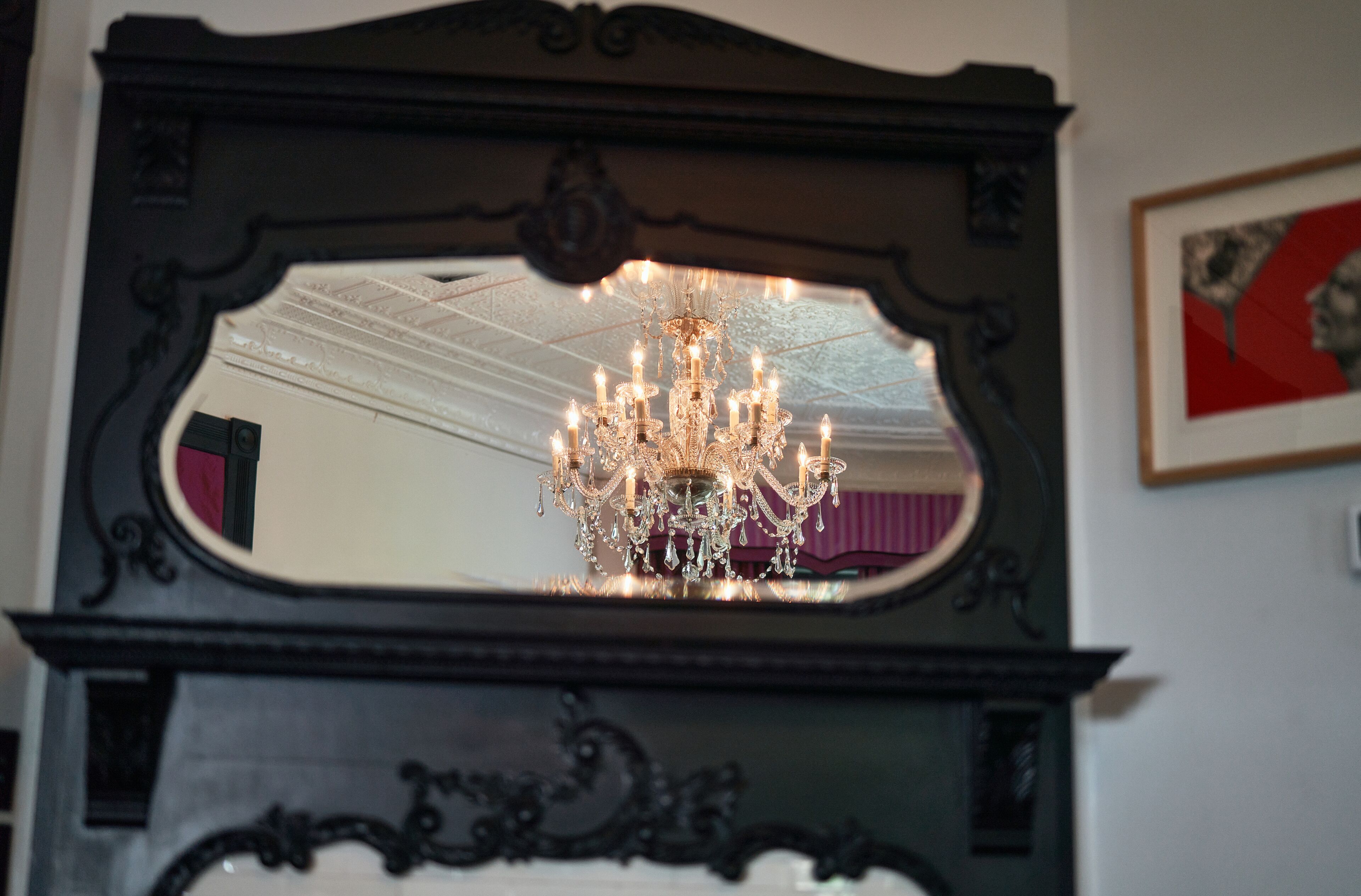 Un mirall amb un candelabrum reflectit en el seu interior, marc negre ornamentat i un quadre al mur.