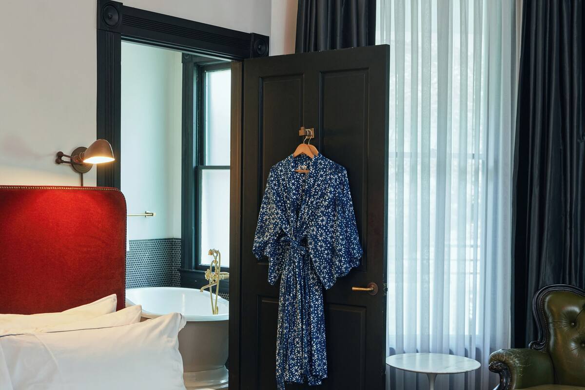 Hotellihuone, jossa on sänky, ovenkahvassa roikkuva kylpytakki, peili, lamppu, tuoli ja ikkunasta avautuva näkymä ulos.