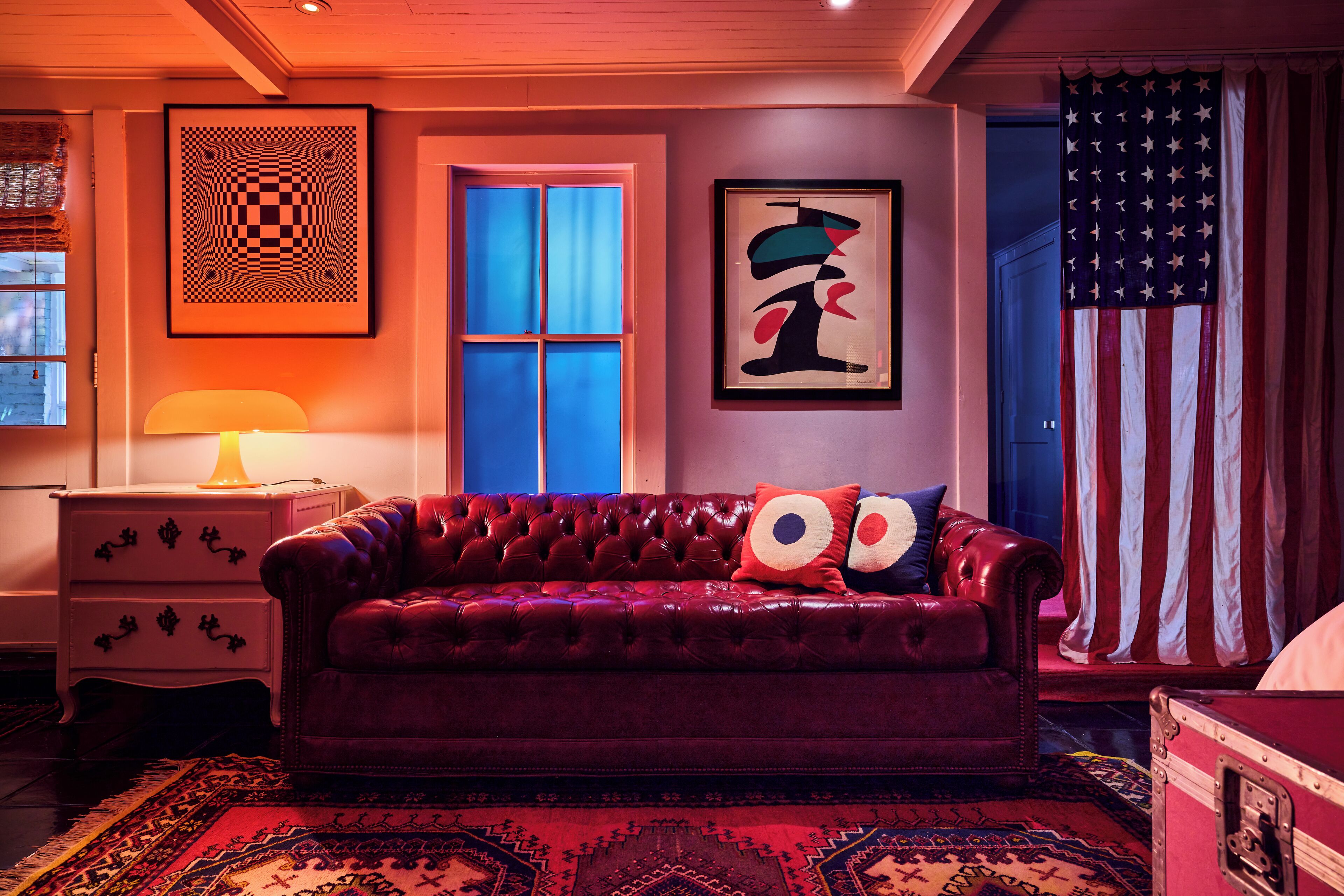 A room with a red velvet sofa, a patterned rug, a framed abstract painting, and an American flag.