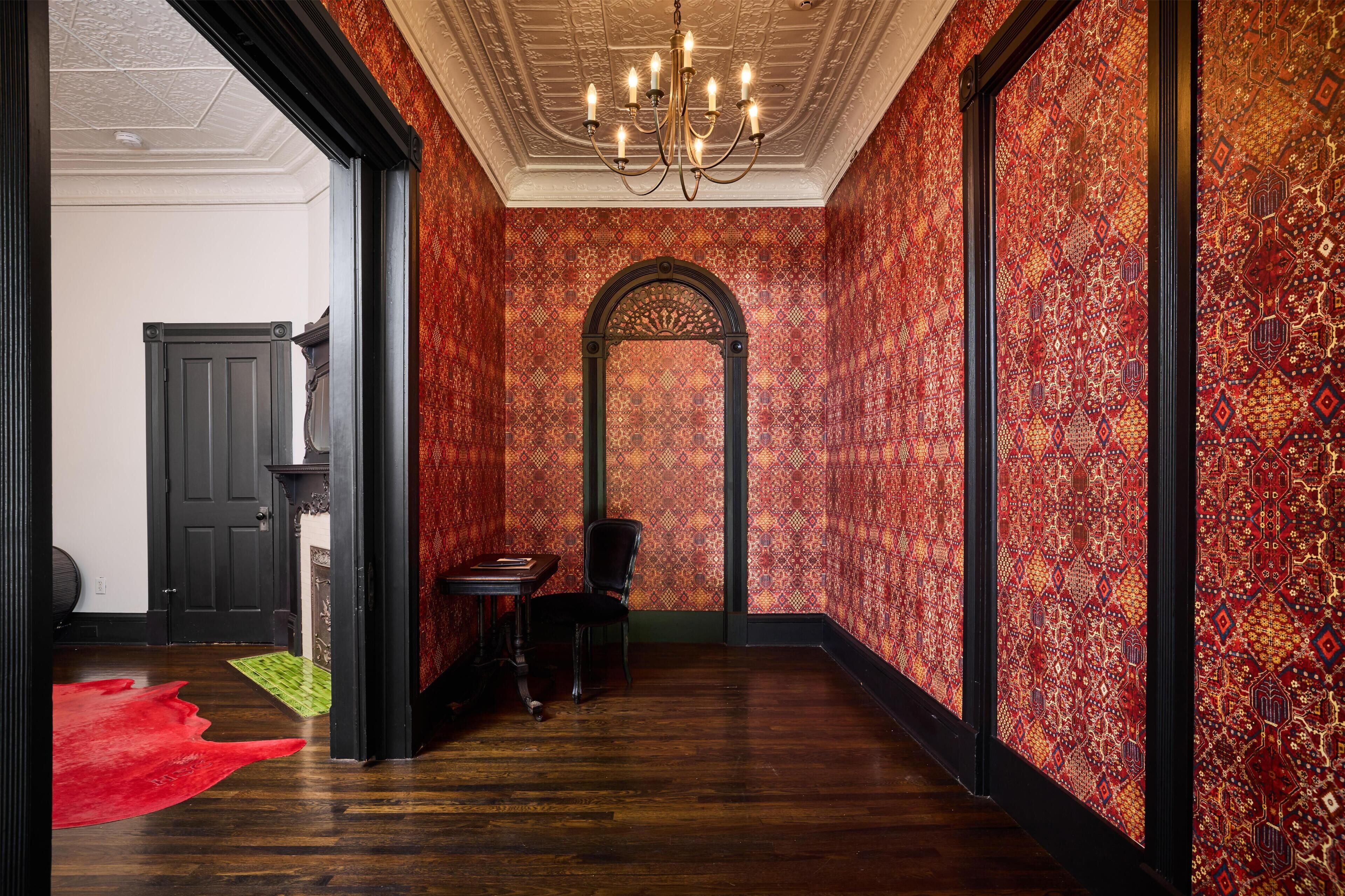 A hallway with patterned wallpaper, a chandelier, a door, and a red rug.
