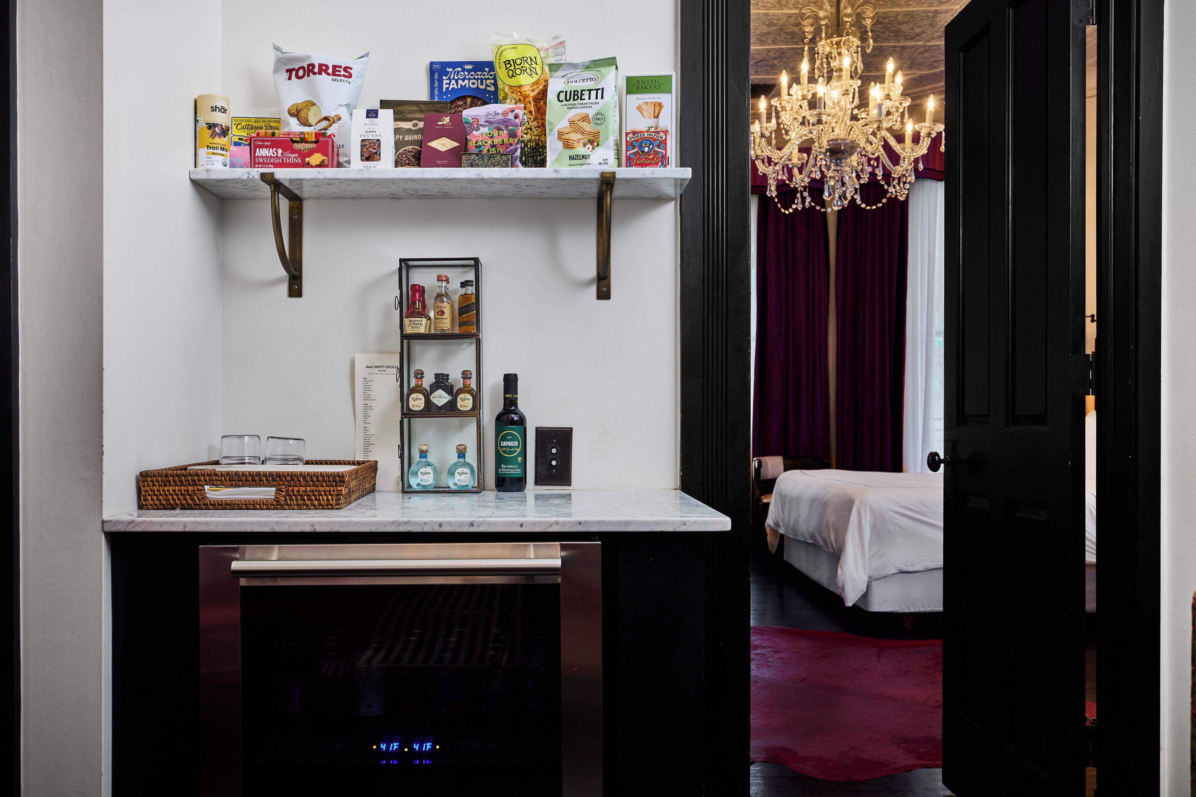 A kitchen area with a countertop, a refrigerator, and a shelf with various food items.