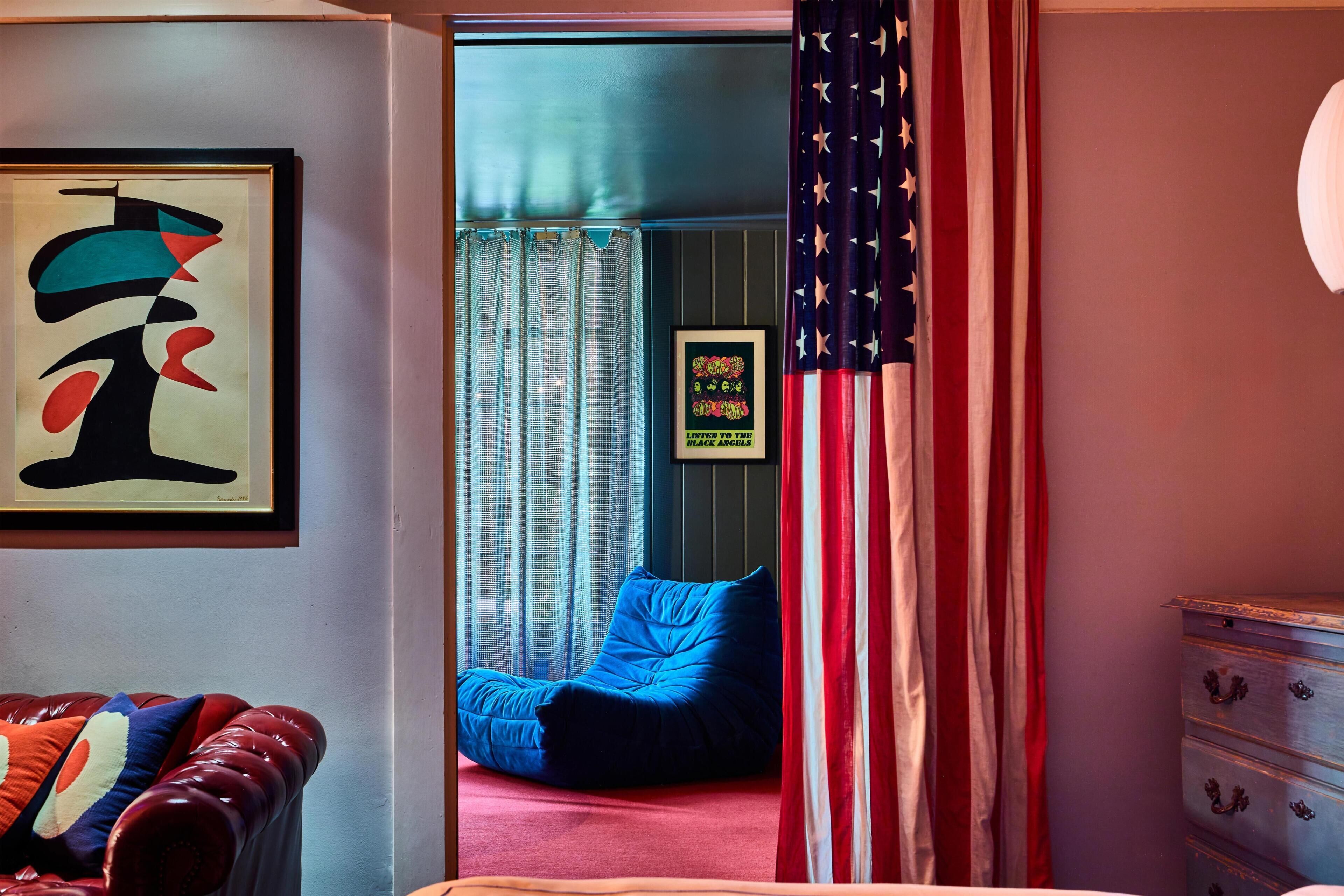 A room with a bed, a red leather chair, a blue bean bag, an American flag, and a framed abstract painting.