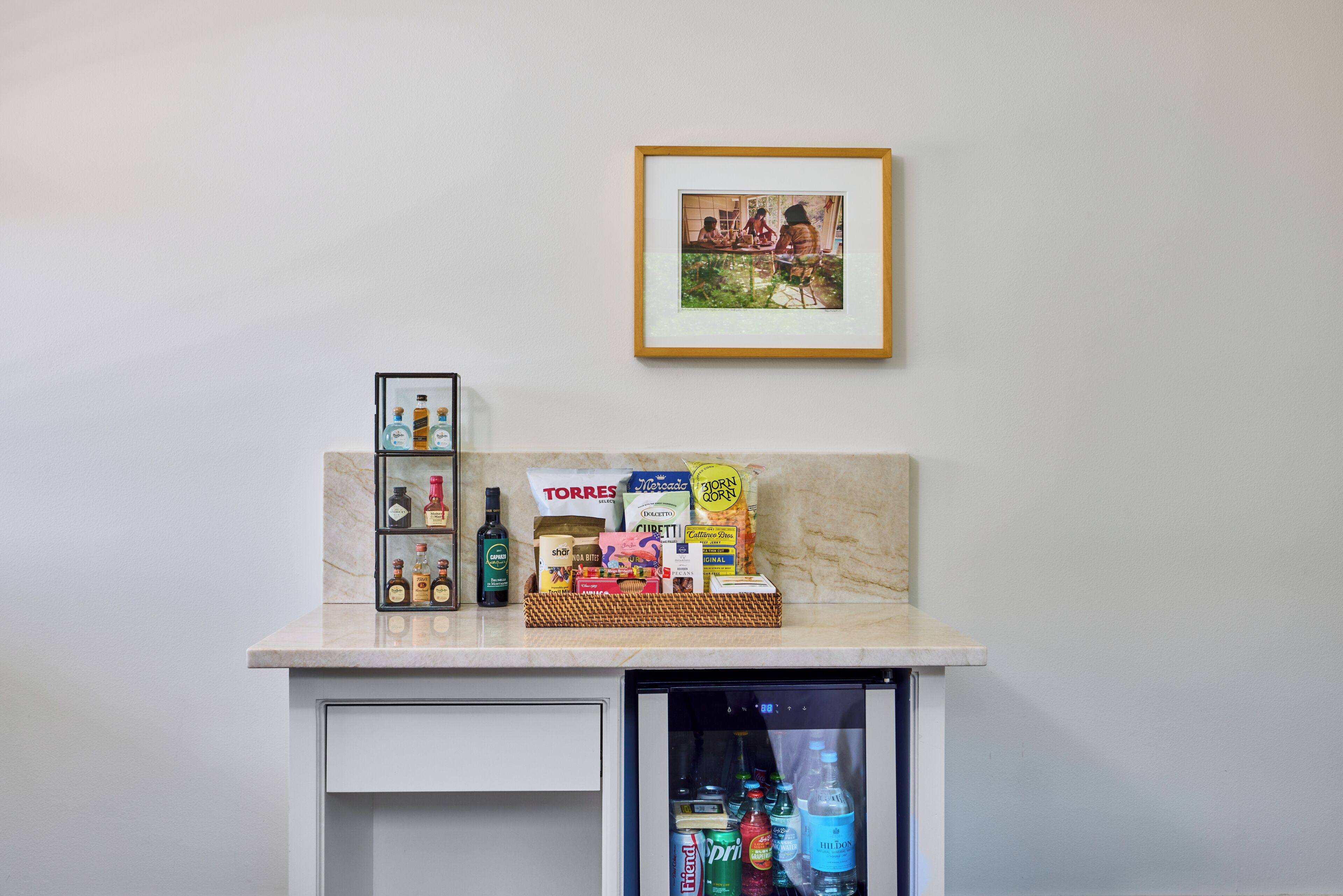 A minibar with a selection of beverages and snacks, a framed picture on the wall, and a small refrigerator below.