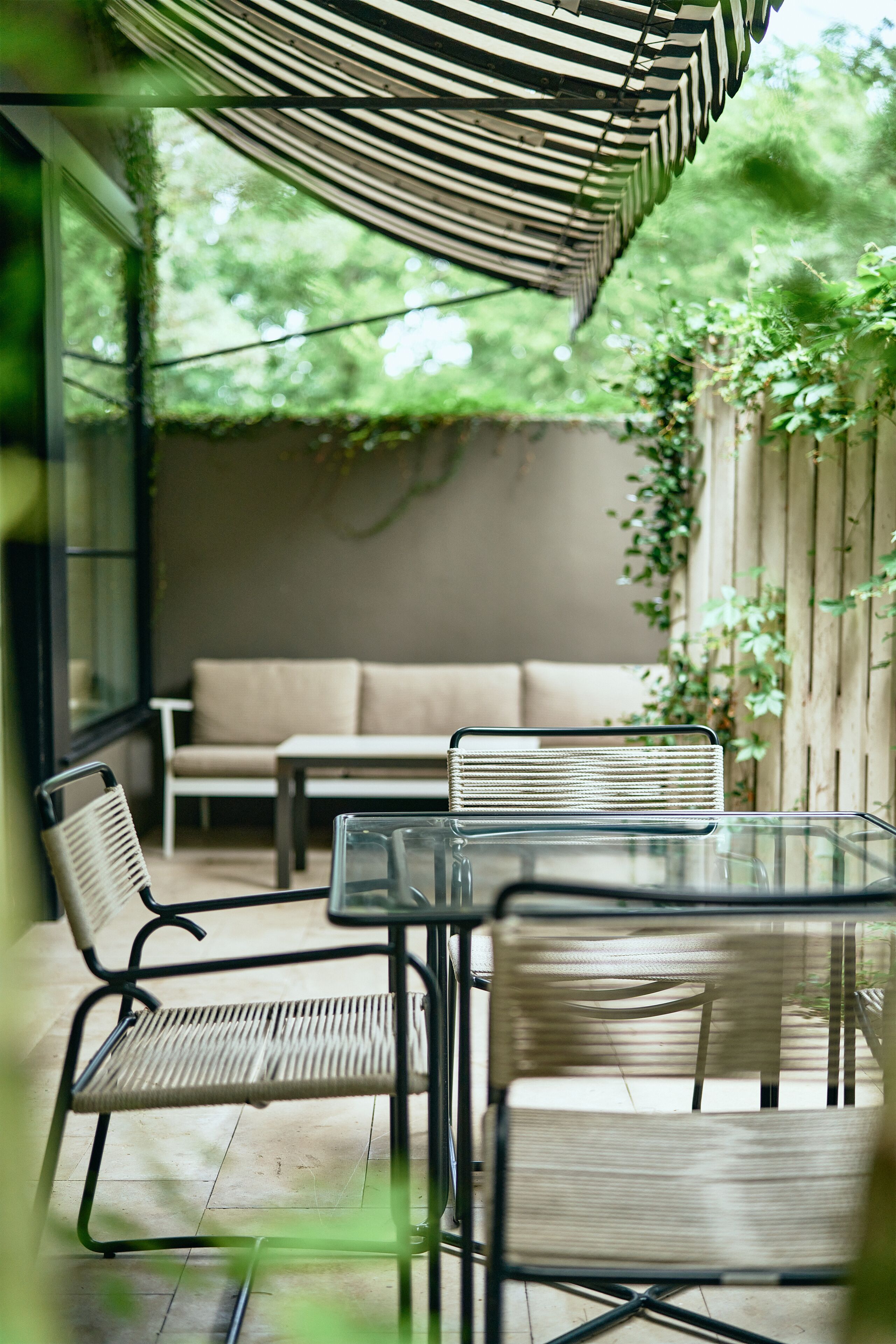 Une terrasse extérieure avec une table en verre et des chaises en osier, un canapé et un auvent à rayures.