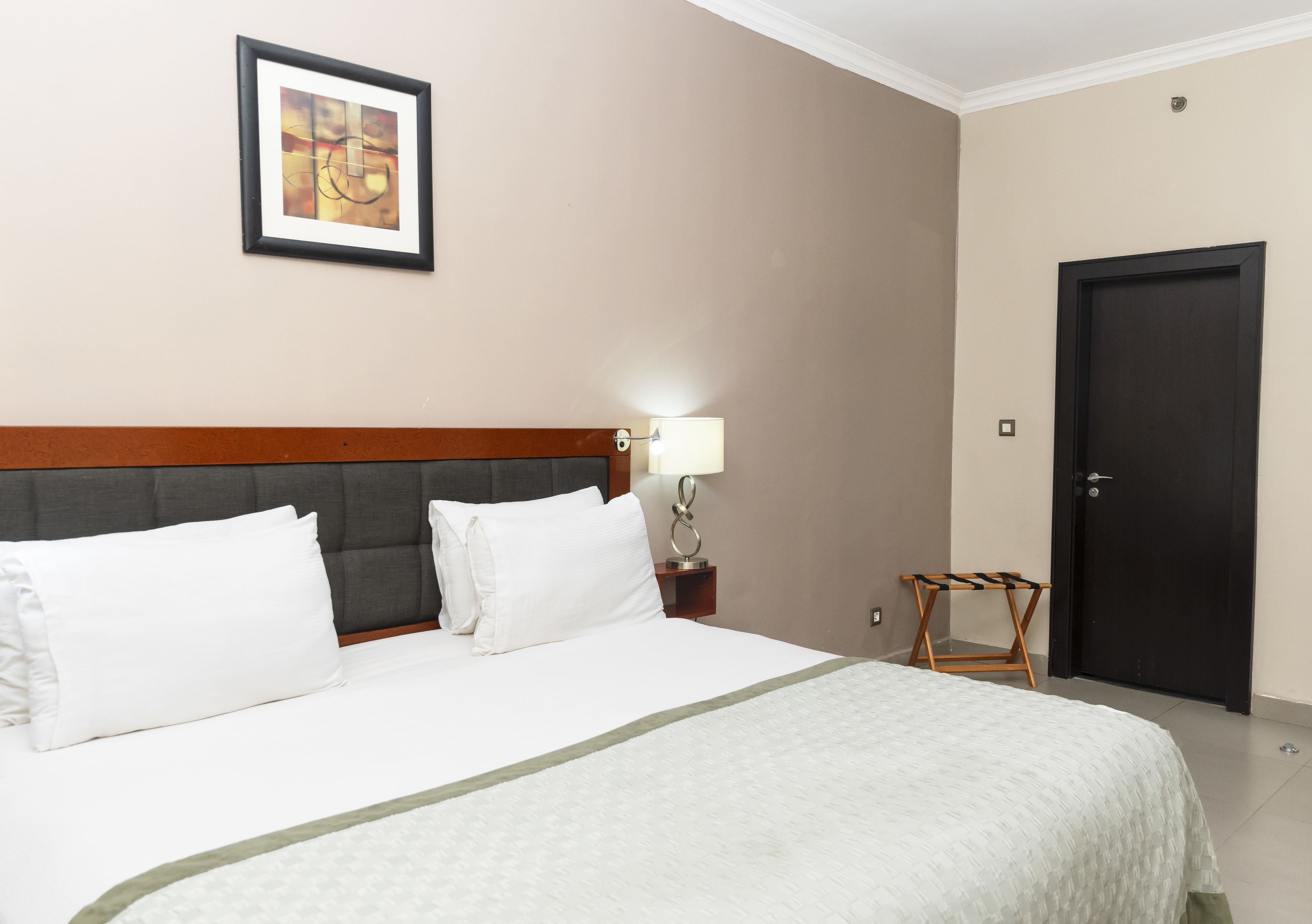 Una habitación de hotel con una cama grande, un cuadro en la pared, una lámpara de noche y una puerta.