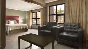 Een hotelkamer met een bed, twee leren fauteuils, een kleine tafel en grote ramen voorzien van gordijnen.