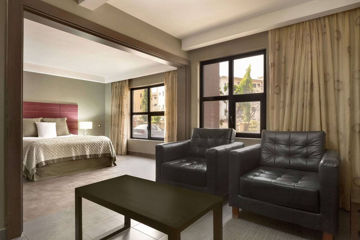 Une chambre d’hôtel avec un lit, deux fauteuils en cuir, une petite table et de grandes fenêtres avec des rideaux.