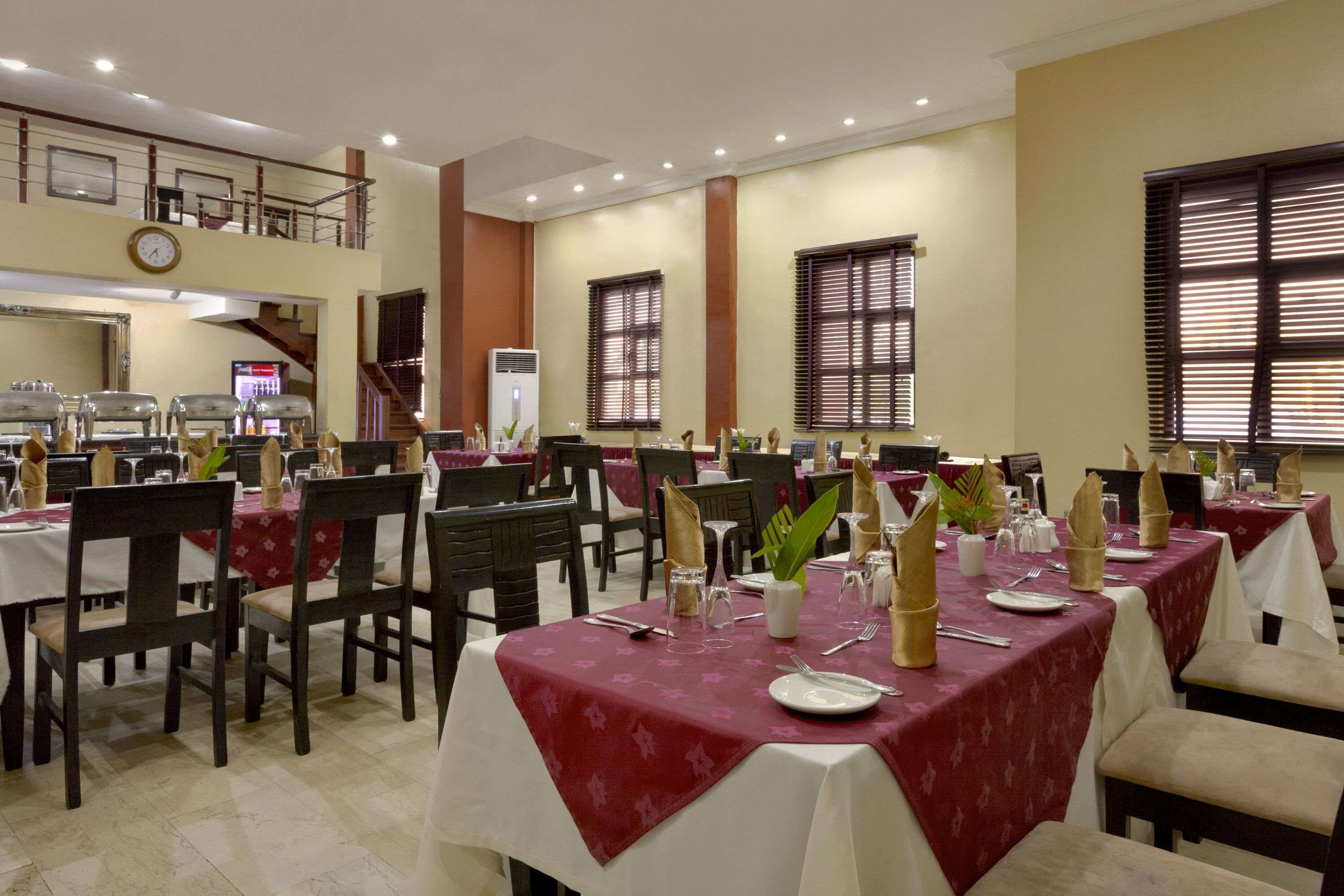 Une salle à manger avec plusieurs tables dressées pour le service, une horloge au mur et un escalier menant à un niveau supérieur.