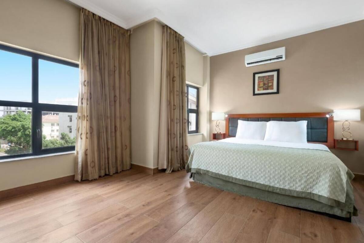 Une chambre d’hôtel dotée d’une grande fenêtre, d’un lit avec une tête de lit, d’une table de chevet avec une lampe et d’un appareil de climatisation fixé au mur.