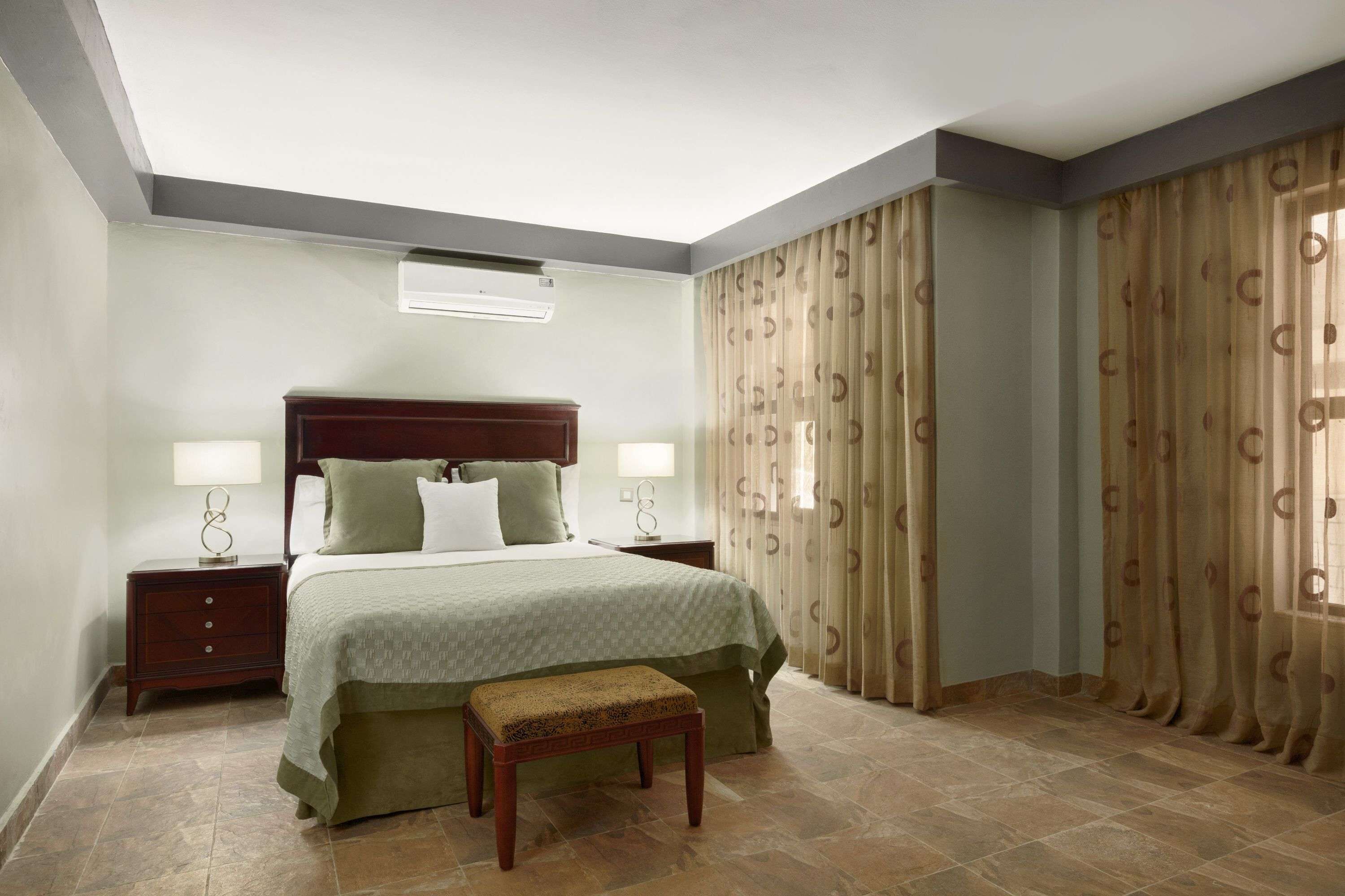 Habitación de hotel con cama, mesitas de noche, un banco, cortinas y un ventilador de techo.