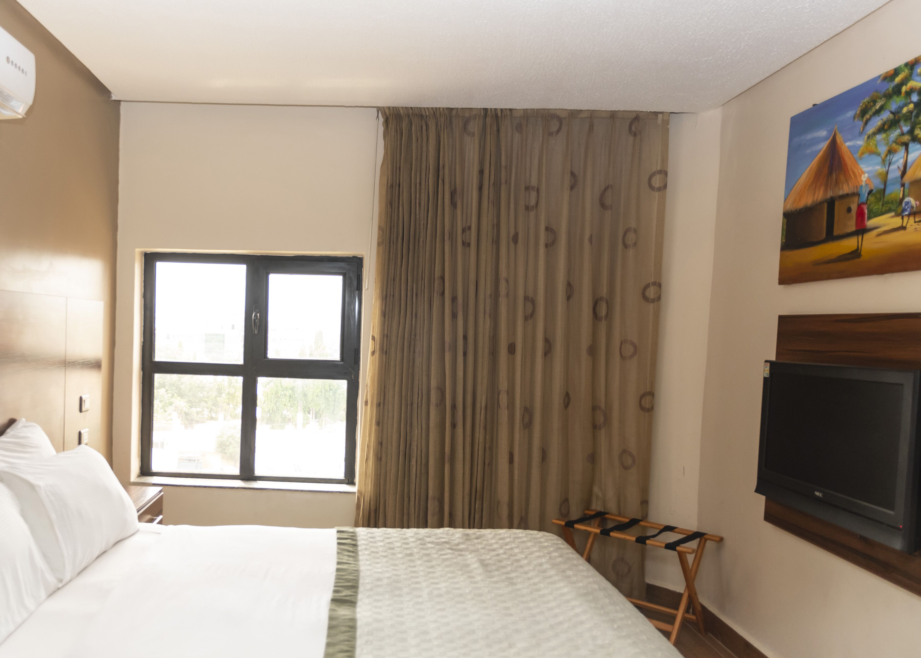 A hotel room with a bed, a window, a television, and a painting on the wall.