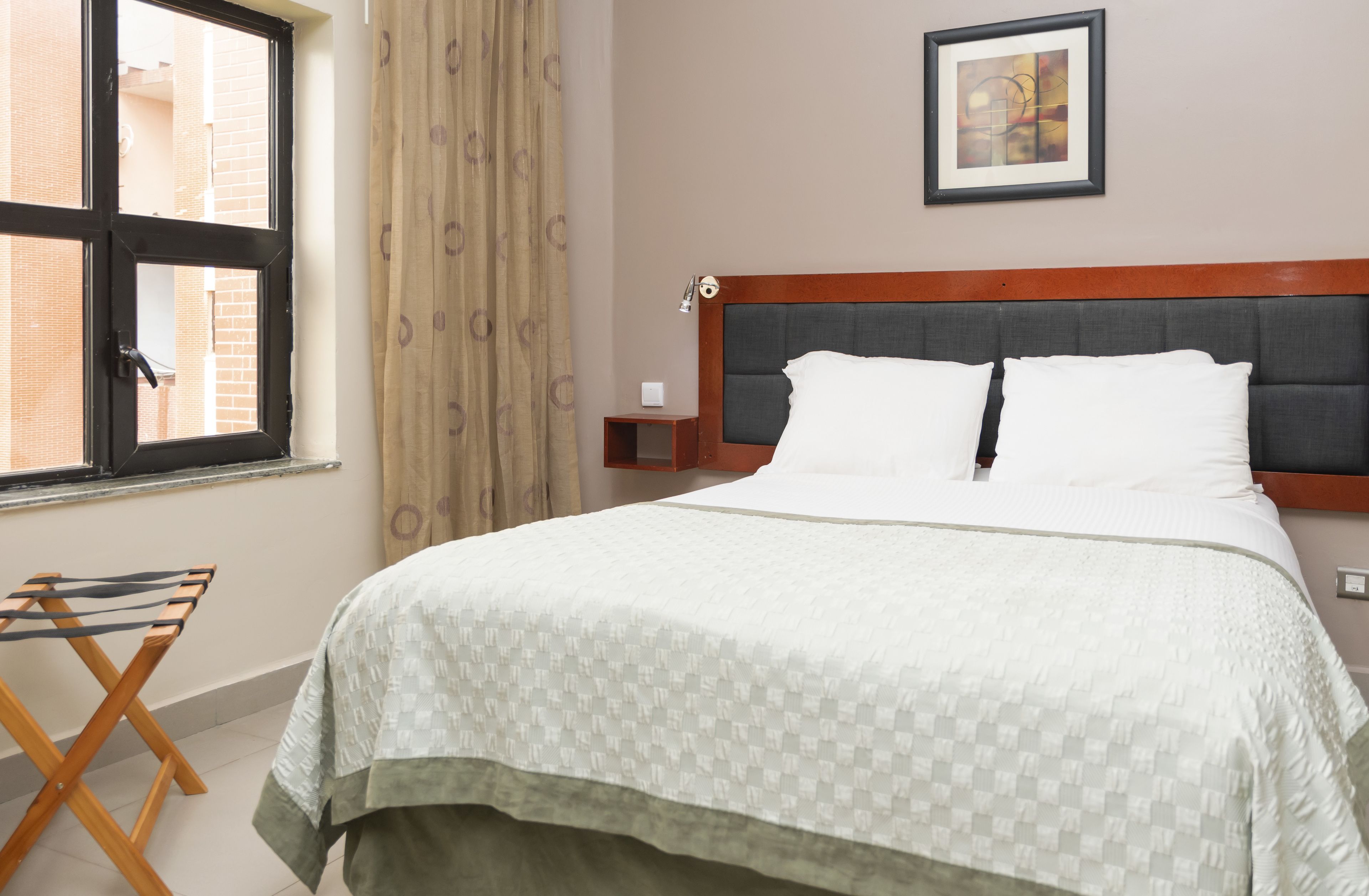 Une chambre d’hôtel avec un lit, une fenêtre avec des rideaux, un tableau accroché au mur et une petite table de chevet.