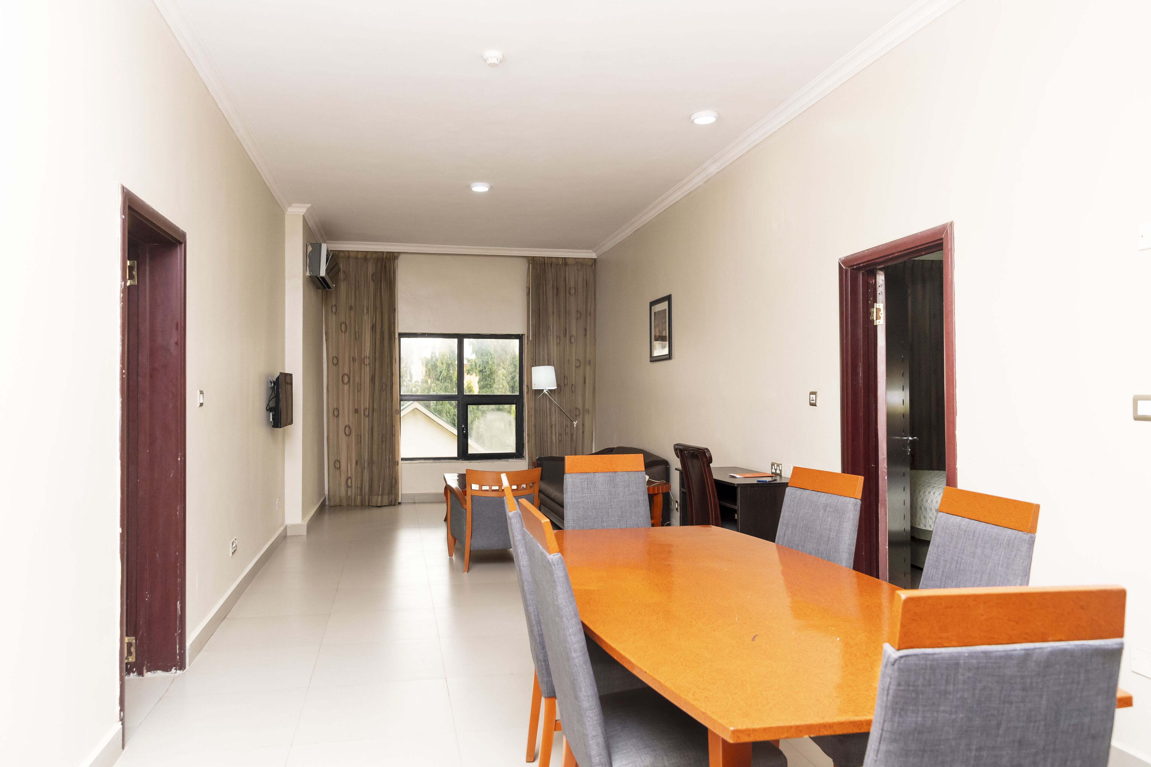 Un grand espace dédié à la salle à manger, avec une longue table en bois et six chaises, une fenêtre avec des rideaux et une porte située à gauche.