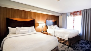 Ein Hotelzimmer mit zwei Betten, Holz-Kopfteilen, einem Nachttisch mit Lampe und einem großen Fenster mit Vorhängen.
