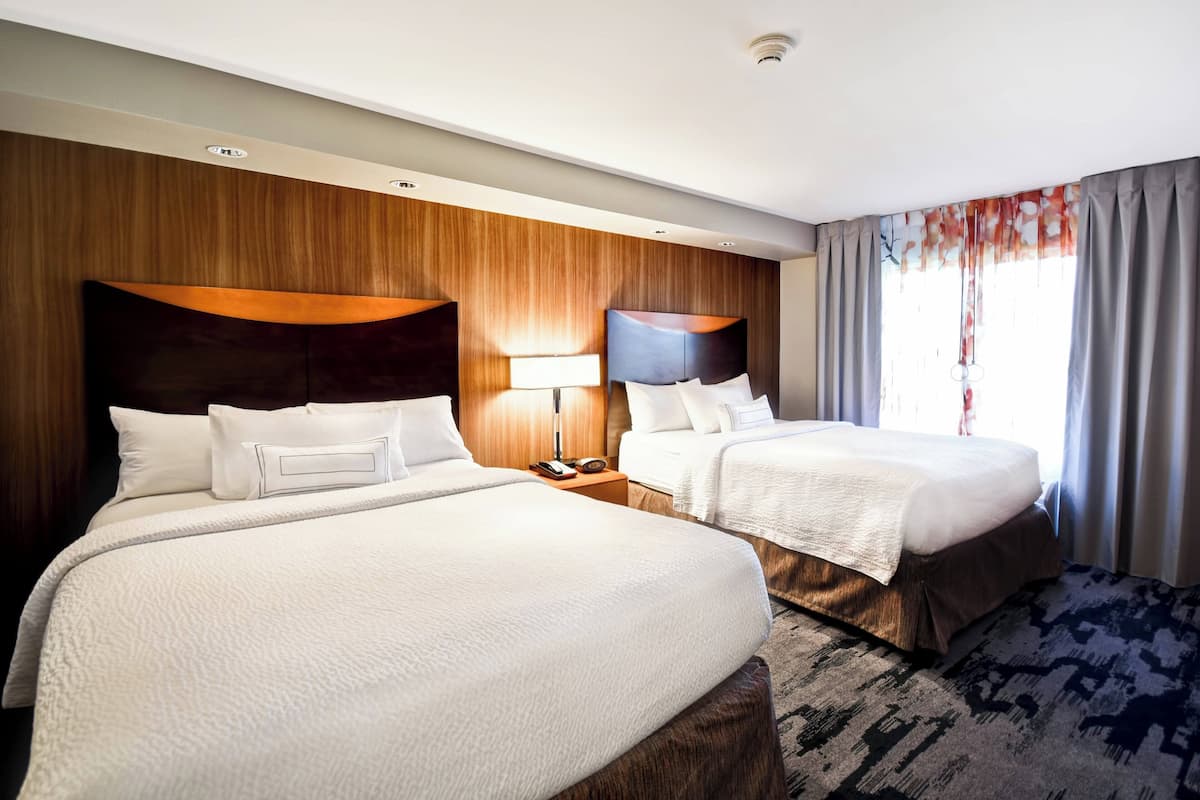 Ein Hotelzimmer mit zwei Betten, Holz-Kopfteilen, einem Nachttisch mit Lampe und einem großen Fenster mit Vorhängen.