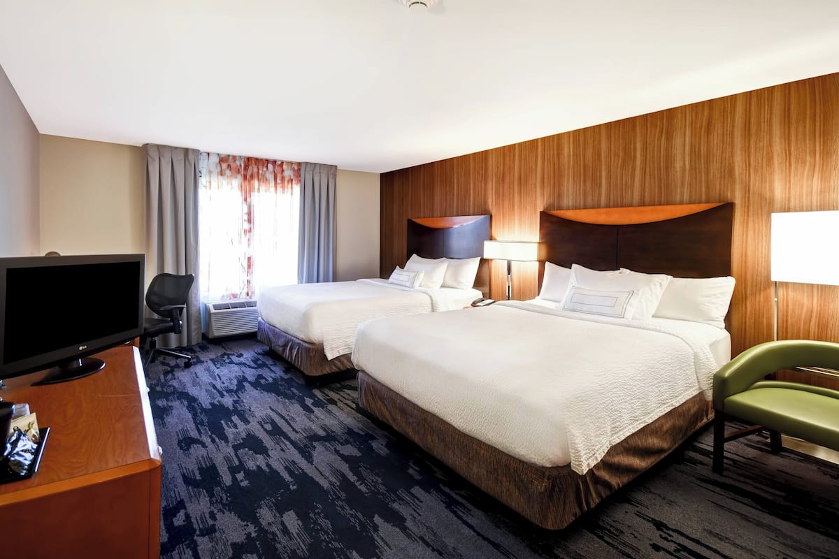 Ein Hotelzimmer mit zwei Betten, einem Schreibtisch mit Fernseher, einem grünen Sessel und einem großen Fenster mit Vorhängen.