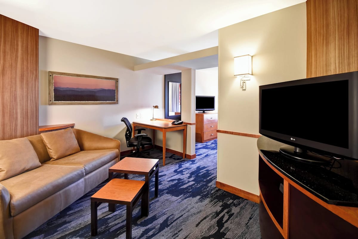 Ein Hotelzimmer mit einer Couch, einem kleinen Tisch, einem Fernseher, einem Schreibtisch mit Stuhl und Teppichboden.