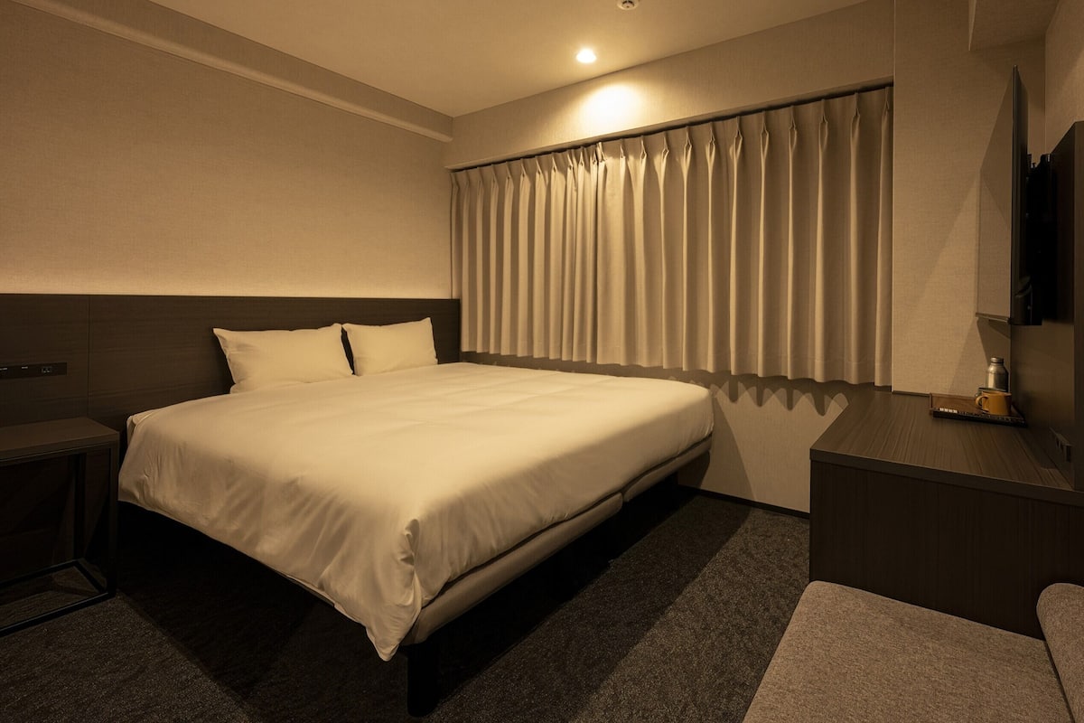 Une chambre d’hôtel avec un lit, une table de chevet, un bureau, un miroir et une fenêtre avec des rideaux.