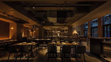 Interior modern d'un restaurant amb diverses taules i cadirees, una cuina al fons i grans finestres a un costat.