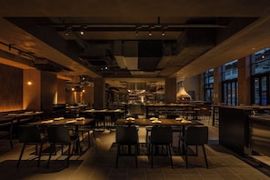 L’intérieur d’un restaurant moderne, avec de nombreuses tables et chaises, un espace cuisine à l’arrière-plan et de grandes fenêtres sur un des côtés.