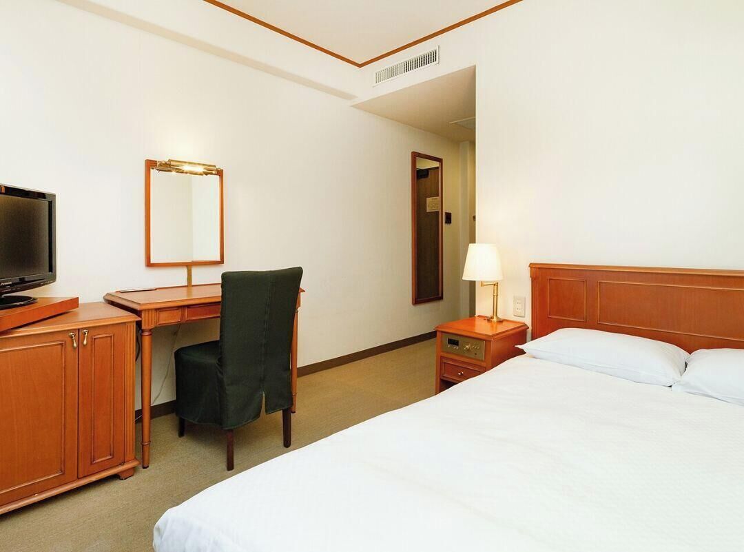 Een hotelkamer met een bed, een bureau met stoel, een televisie, een spiegel en een bedlamp.