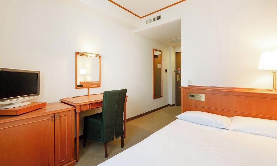 Een hotelkamer met een bed, een bureau met stoel, een televisie, een spiegel en een lamp.