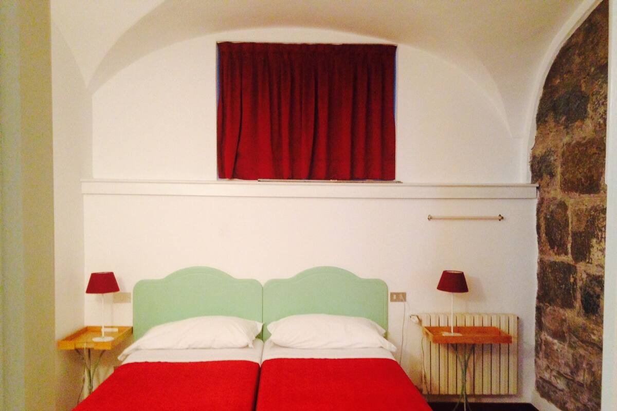 A room with a bed, two bedside tables, a window with red curtains, a radiator, and a stone wall.