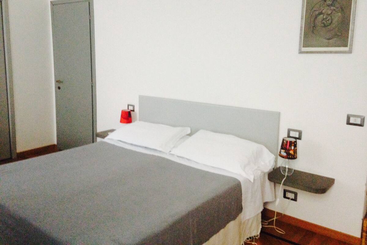A simple, clean room with a bed, a bedside table, a red object on the table, and a framed picture on the wall.
