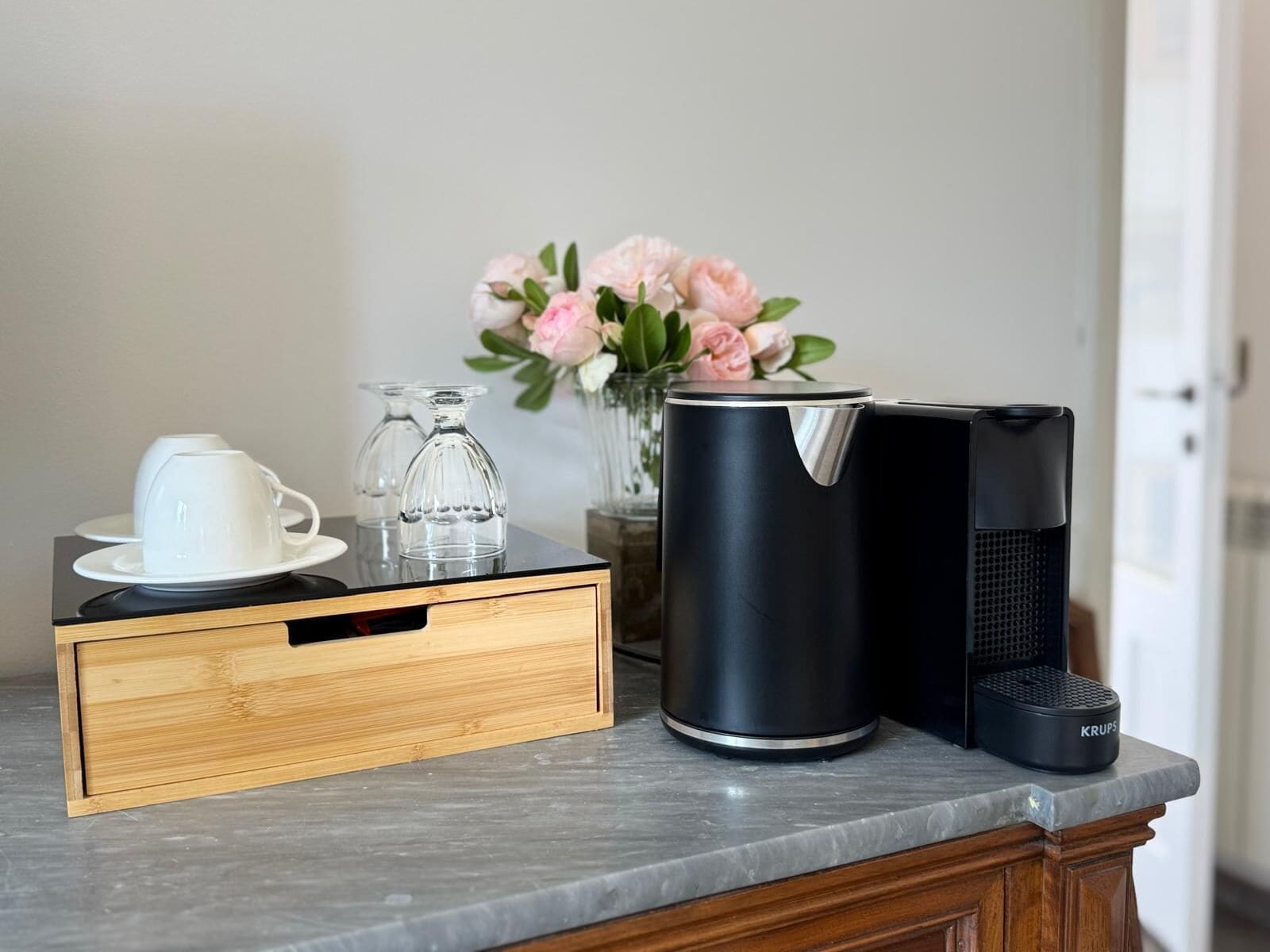 Eine Küchenarbeitsplatte mit einer Kaffeemaschine, einem Glasbehälter, einer weißen Tasse und einer Vase mit rosa Blumen.