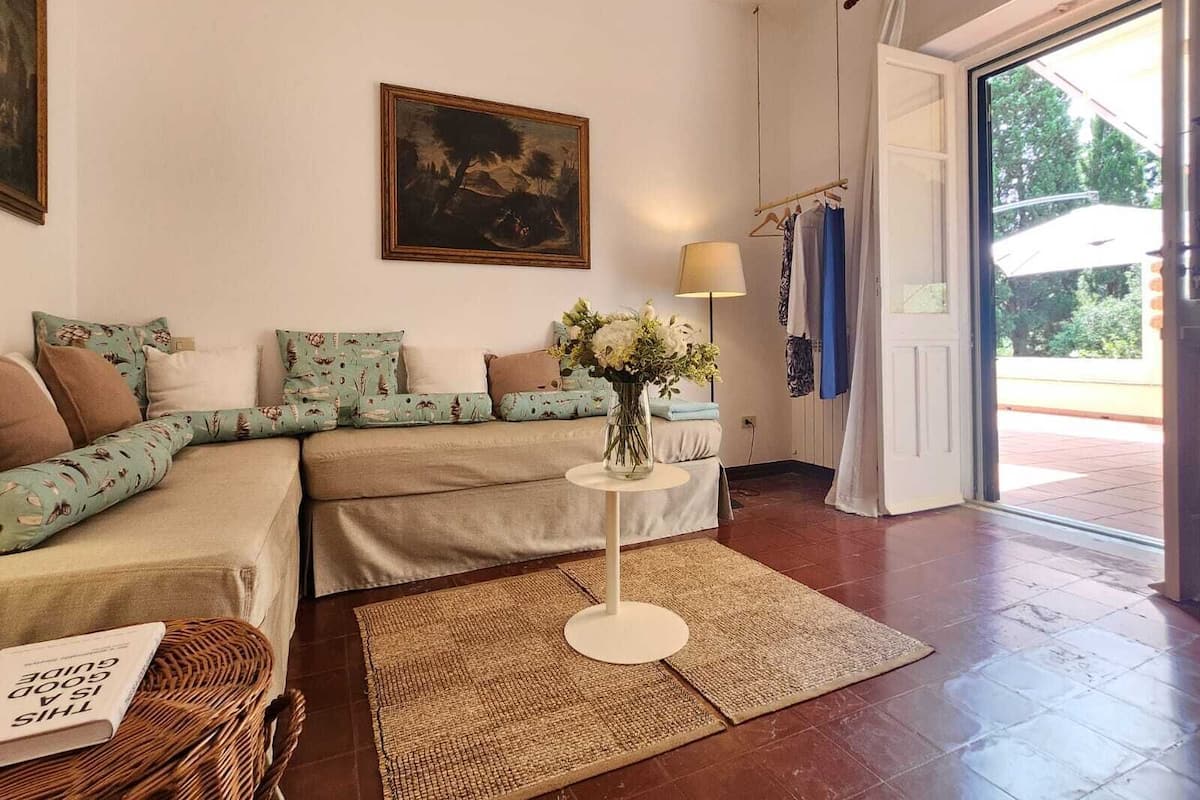 A cozy living room with a sofa, a small table with a lamp, a painting on the wall, and a view through a door.
