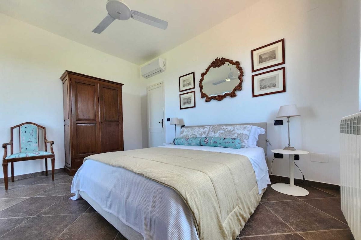 A bedroom with a bed, a wooden wardrobe, a chair, a mirror, and framed pictures on the wall.