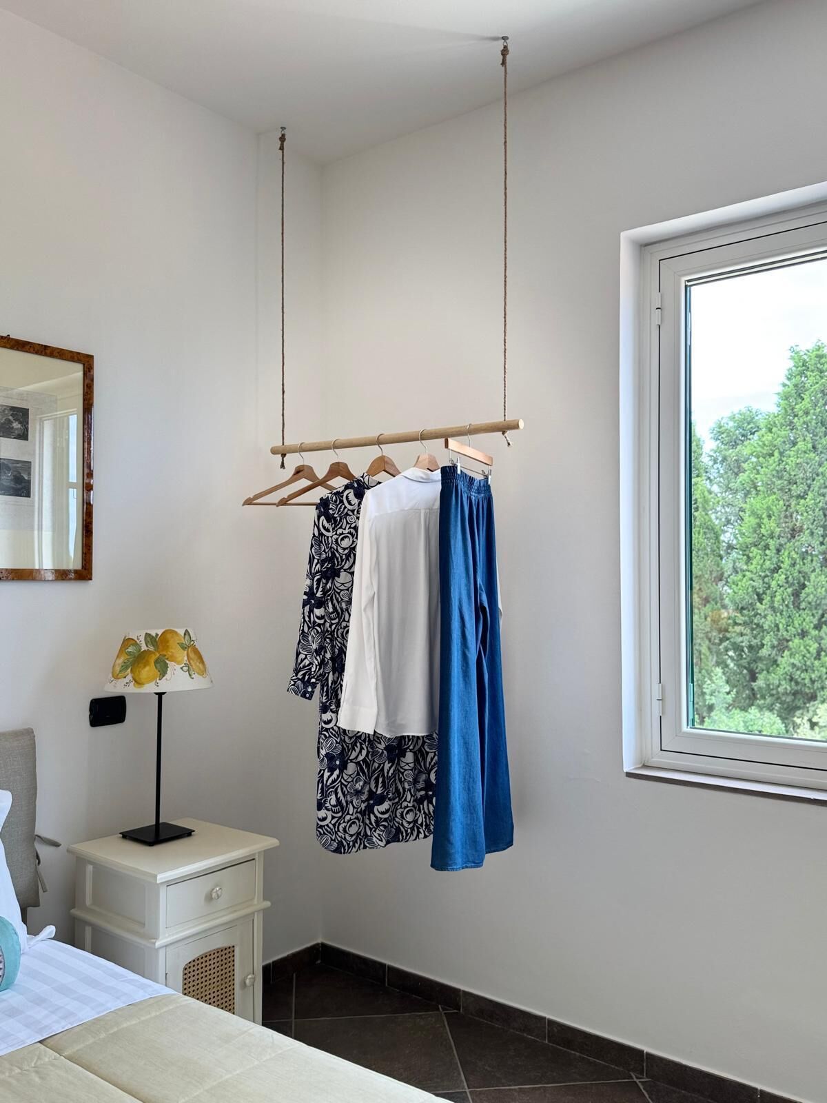 Quarto com cama, mesa de cabeceira, cabideiro, luminária e janela com vista para árvores.