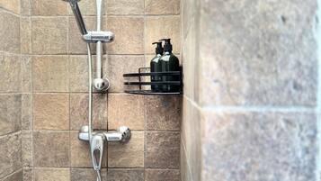 A shower with a handheld showerhead and a wall-mounted faucet, a soap dispenser, and a small shelf with toiletries.